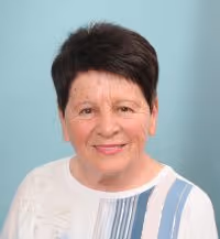 Portrait of a smiling elderly woman with short dark hair wearing a white top with blue and gray stripes on a light blue background.