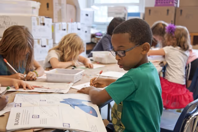 Children seated at a classroom table engaged in writing and drawing activities.