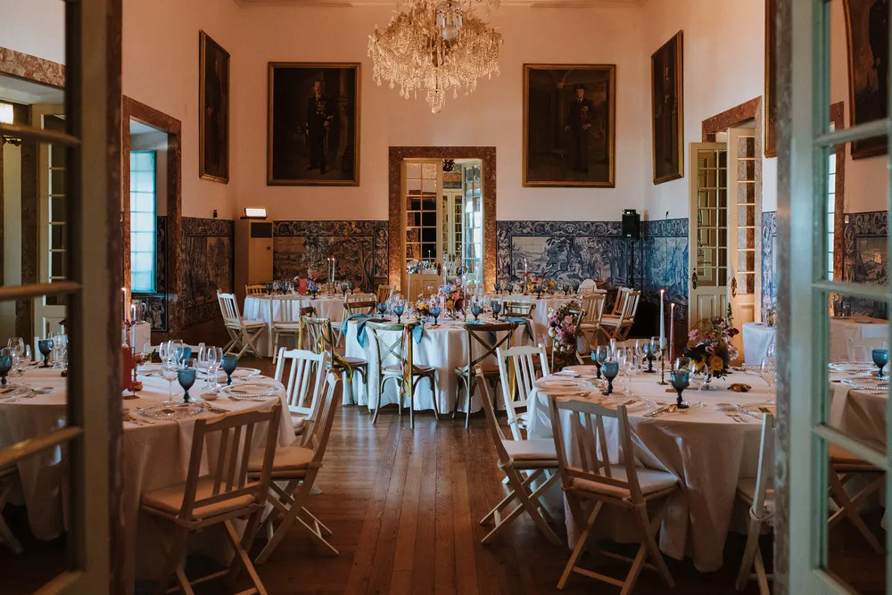 Elegant dining room with round tables, white chairs, floral centerpieces, and a crystal chandelier.