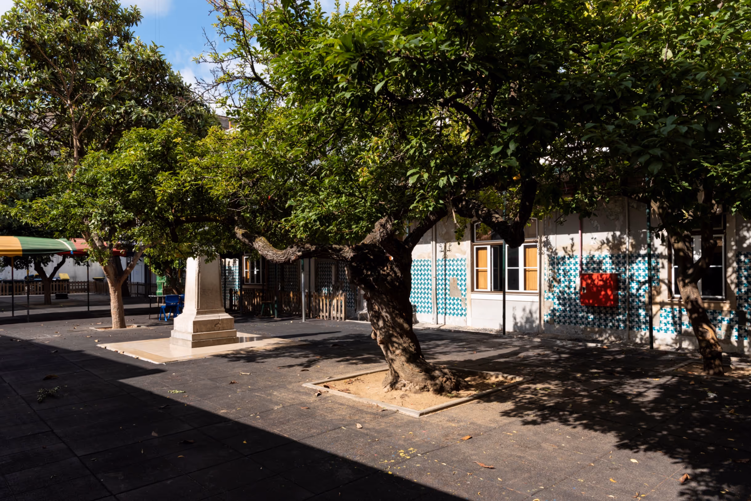 Shaded courtyard with trees, a stone monument, and a tiled building wall with windows.