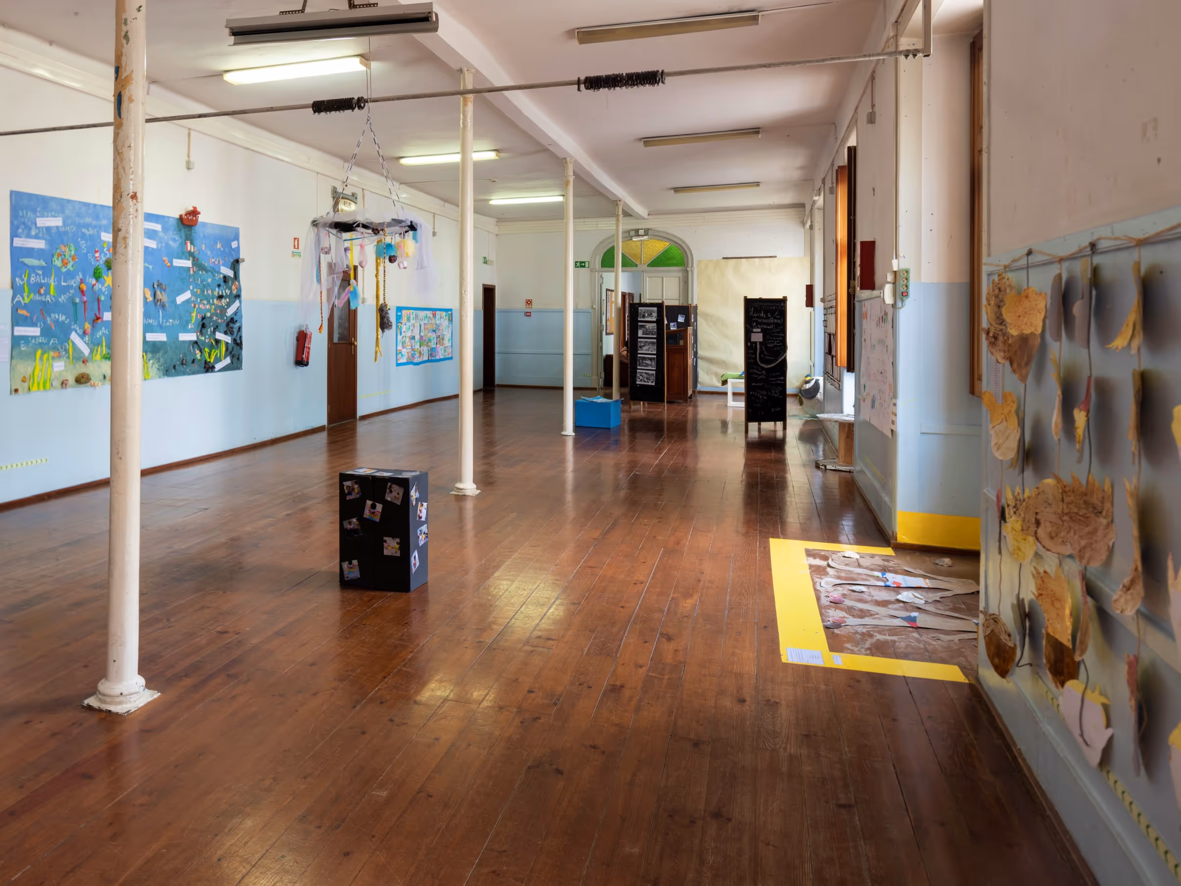 Empty room with wooden floor, white pillars, children's artwork on walls, and hanging craft decorations.