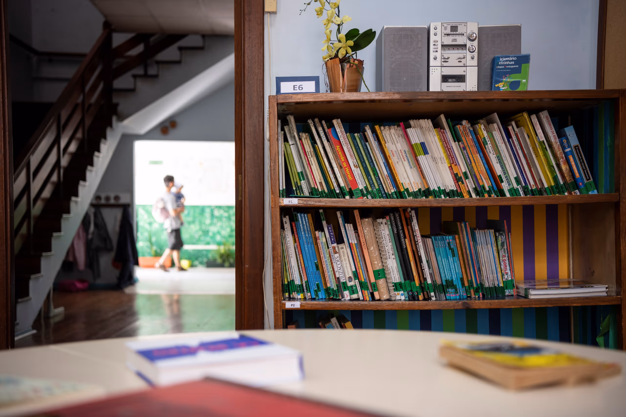 Bookshelf filled with books and a stereo, with blurred person carrying child in background hallway.