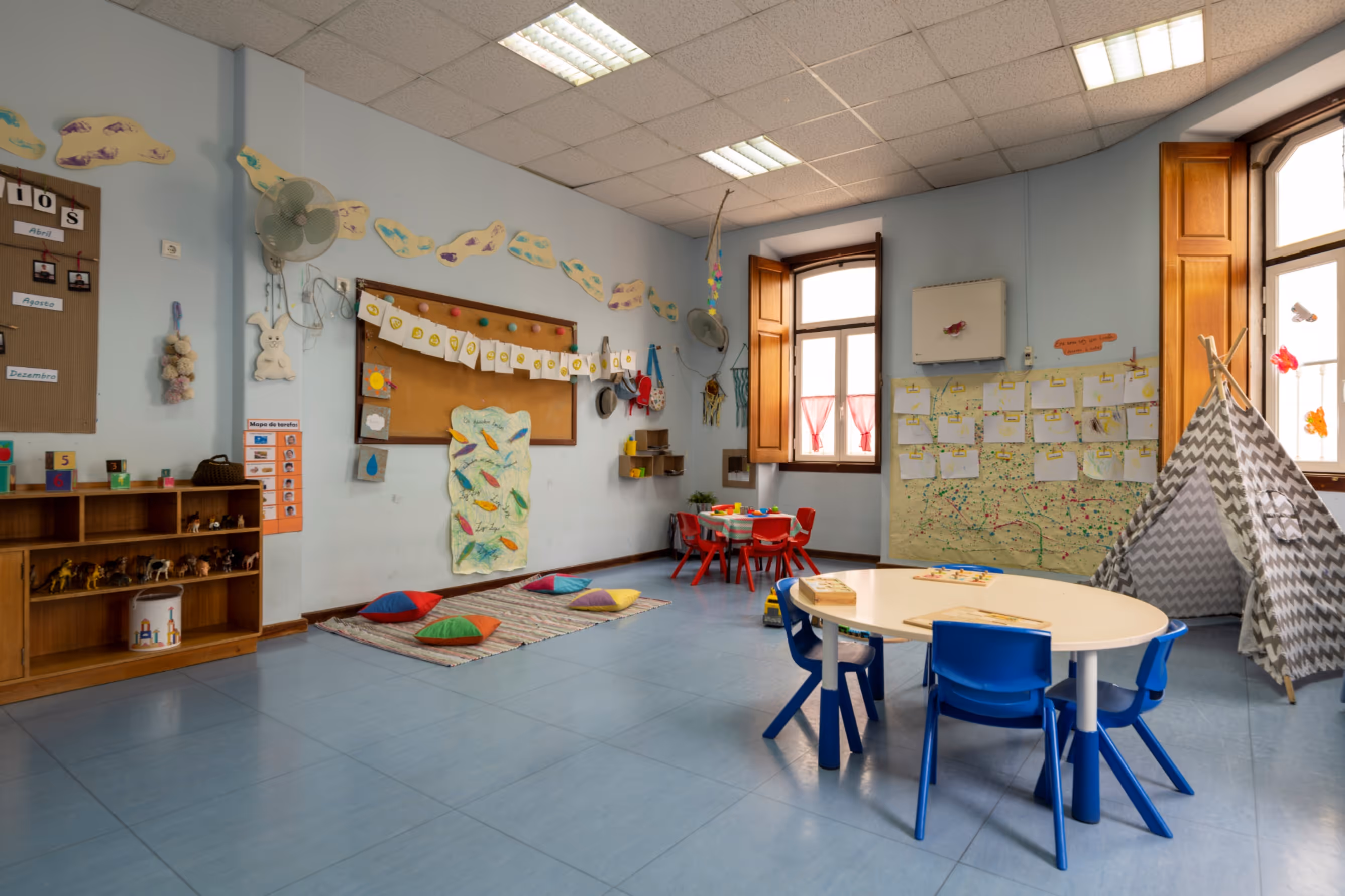 Colorful preschool classroom with chairs, tables, cushions, a play tent, and educational decorations on walls.