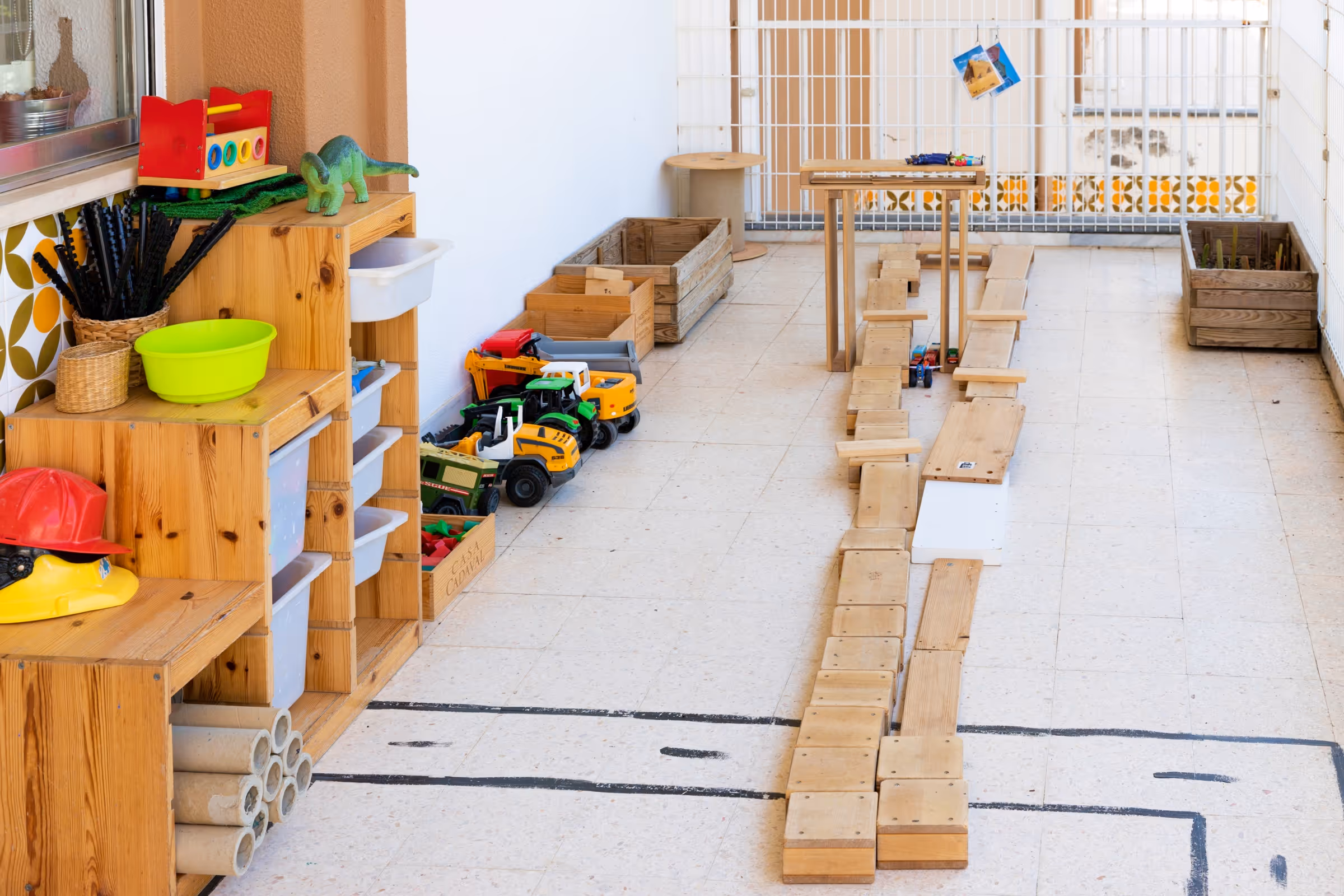 Play area with wooden shelves, toy trucks, dinosaur, helmets, and a wooden race track on the floor.