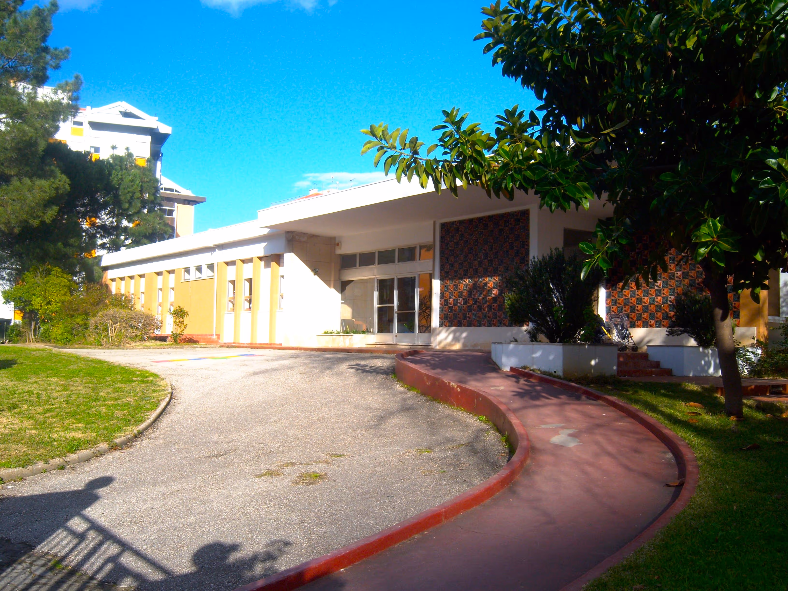Single-story building with a curved red pathway, trees, and clear blue sky on a sunny day.