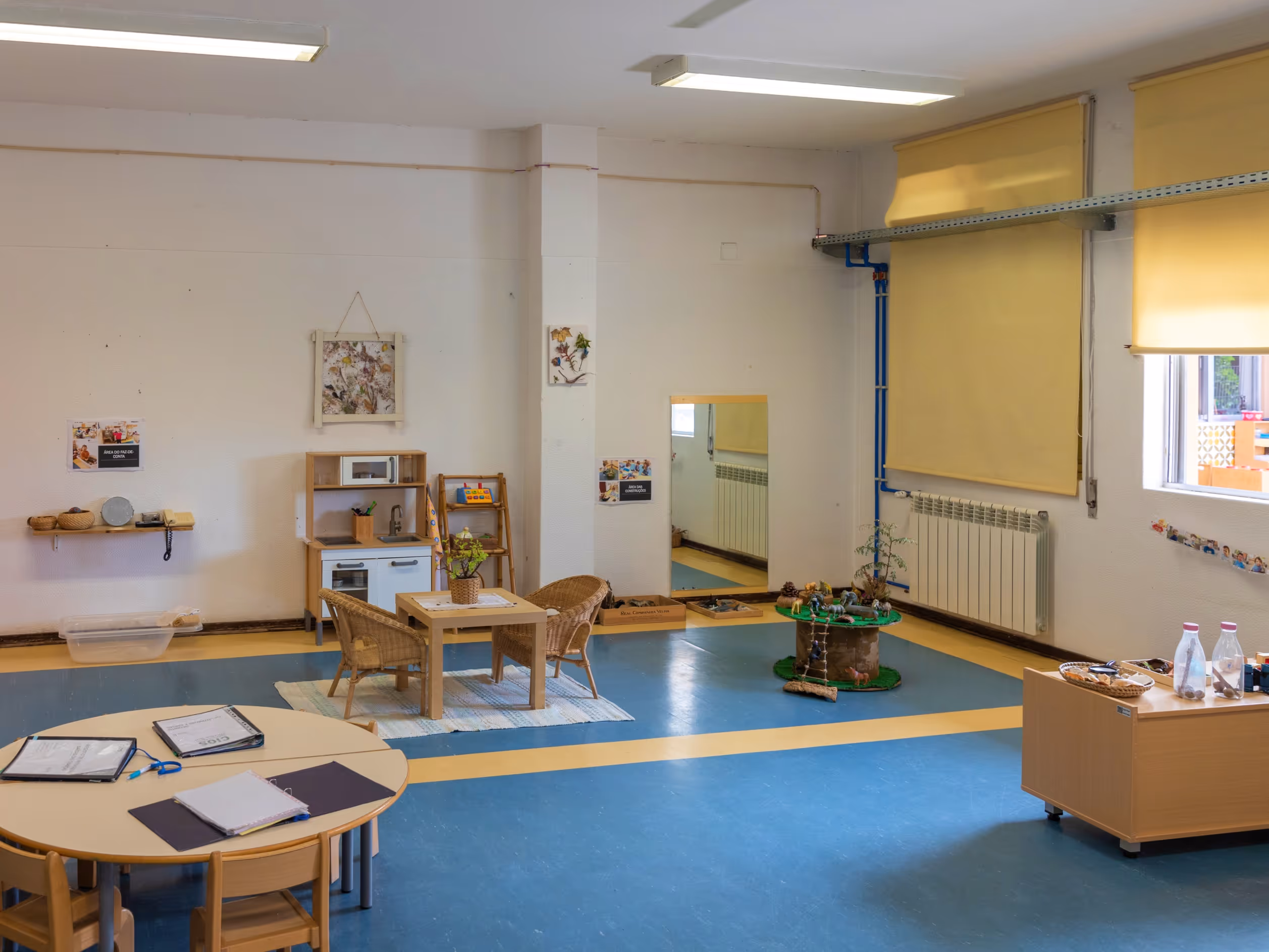 Bright classroom with small tables, chairs, play kitchen, mirror, and educational toys on blue and yellow floor.