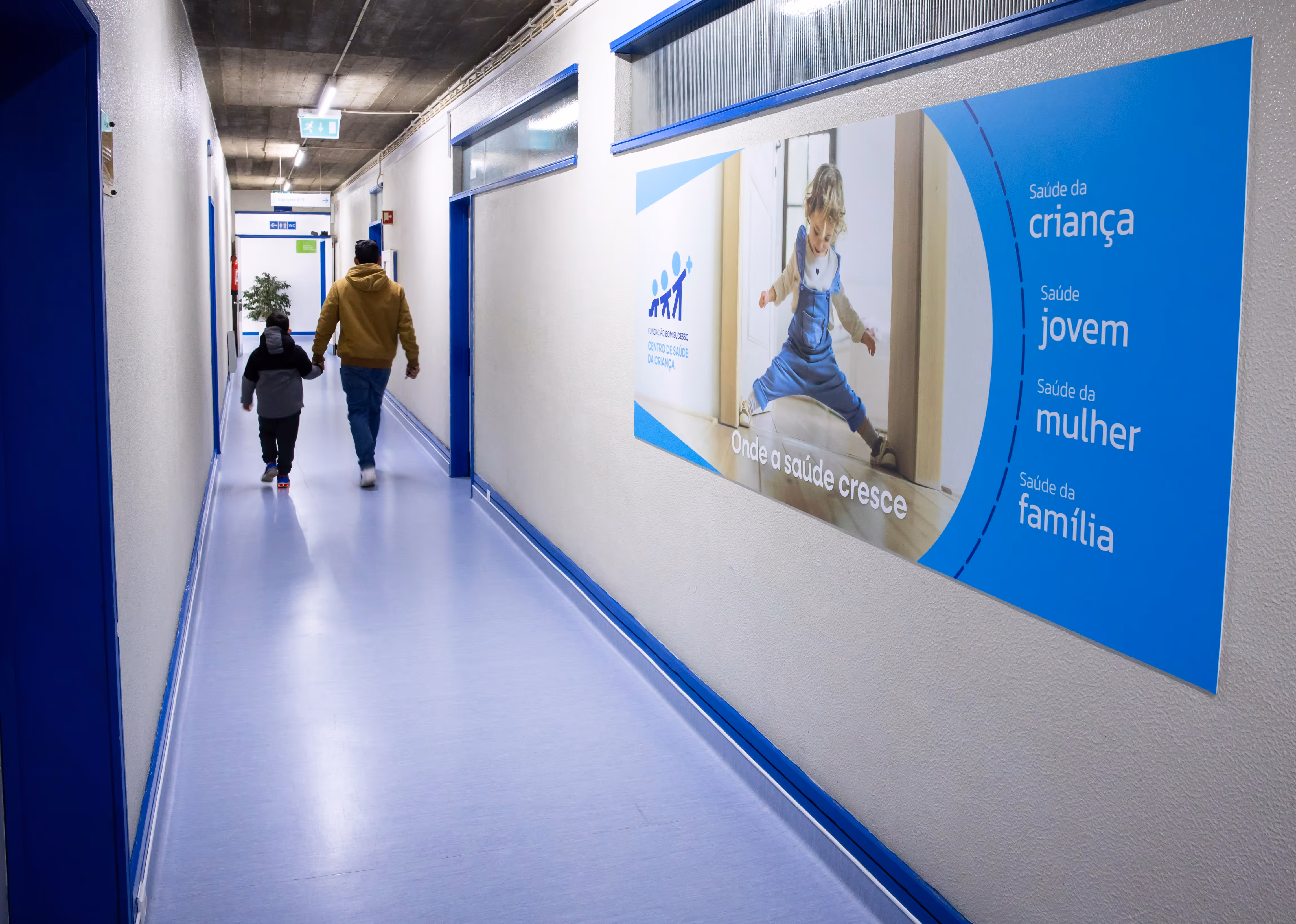 Man holding a child's hand walking down a hospital corridor with a health care poster on the wall.