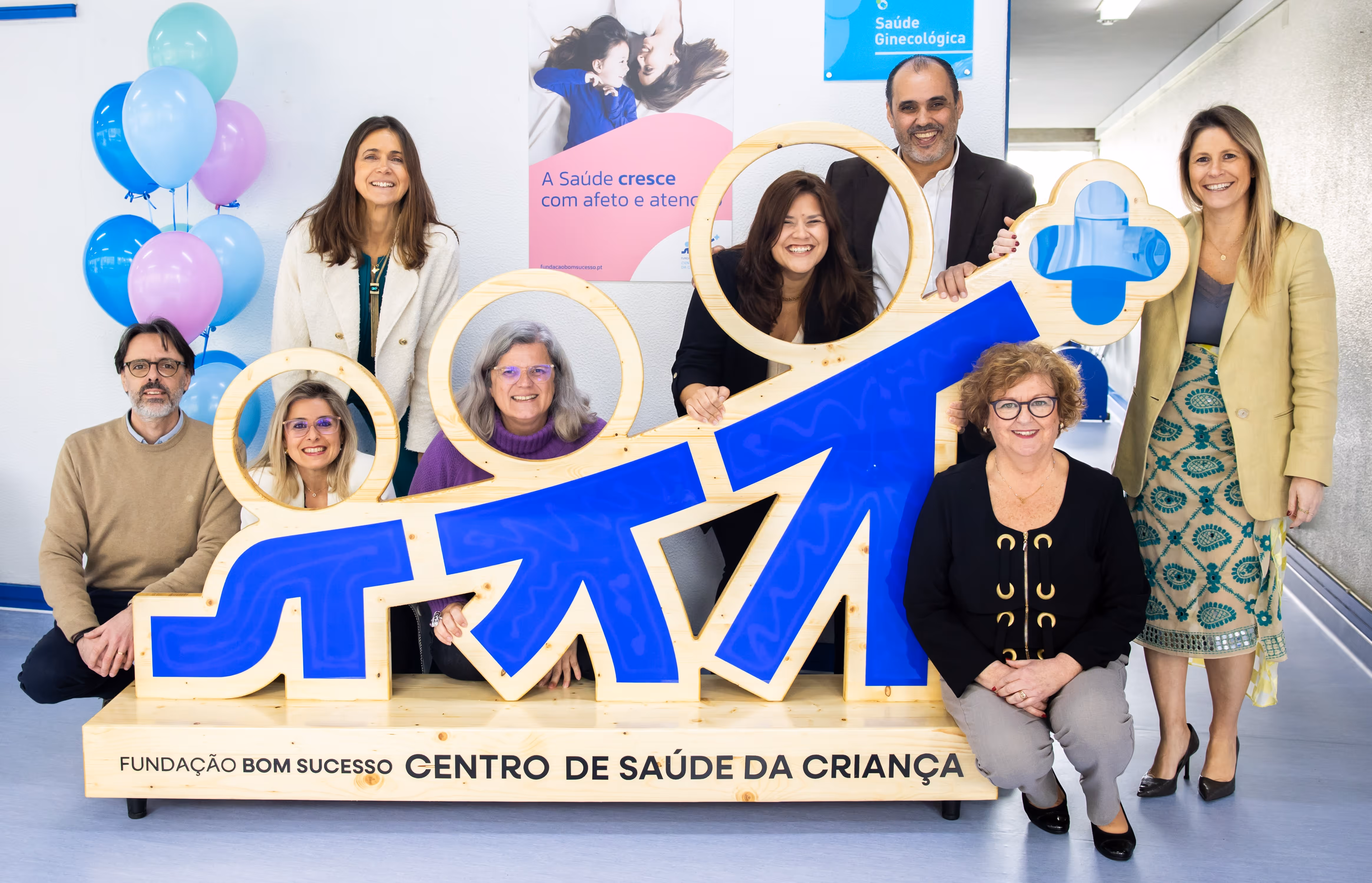 Eight smiling people posing around a large blue and wood Centro de Saúde da Criança sign with balloons nearby.