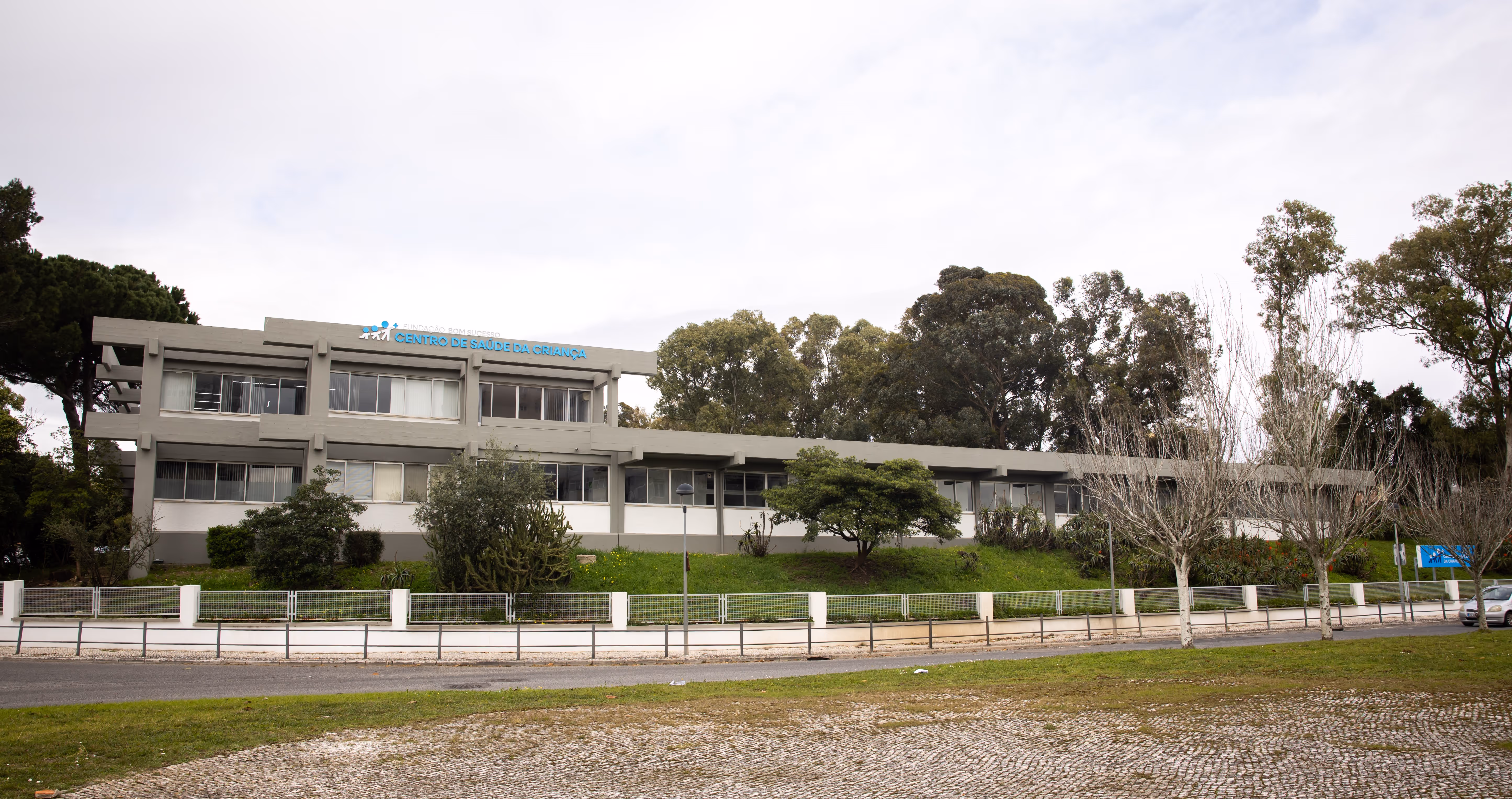 Modern two-story health center building with trees and a road in front under a cloudy sky.