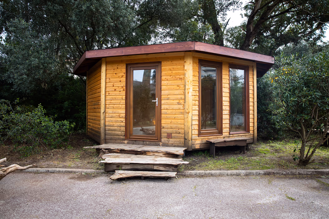 Small wooden cabin with large windows and rustic wooden steps surrounded by trees and greenery.