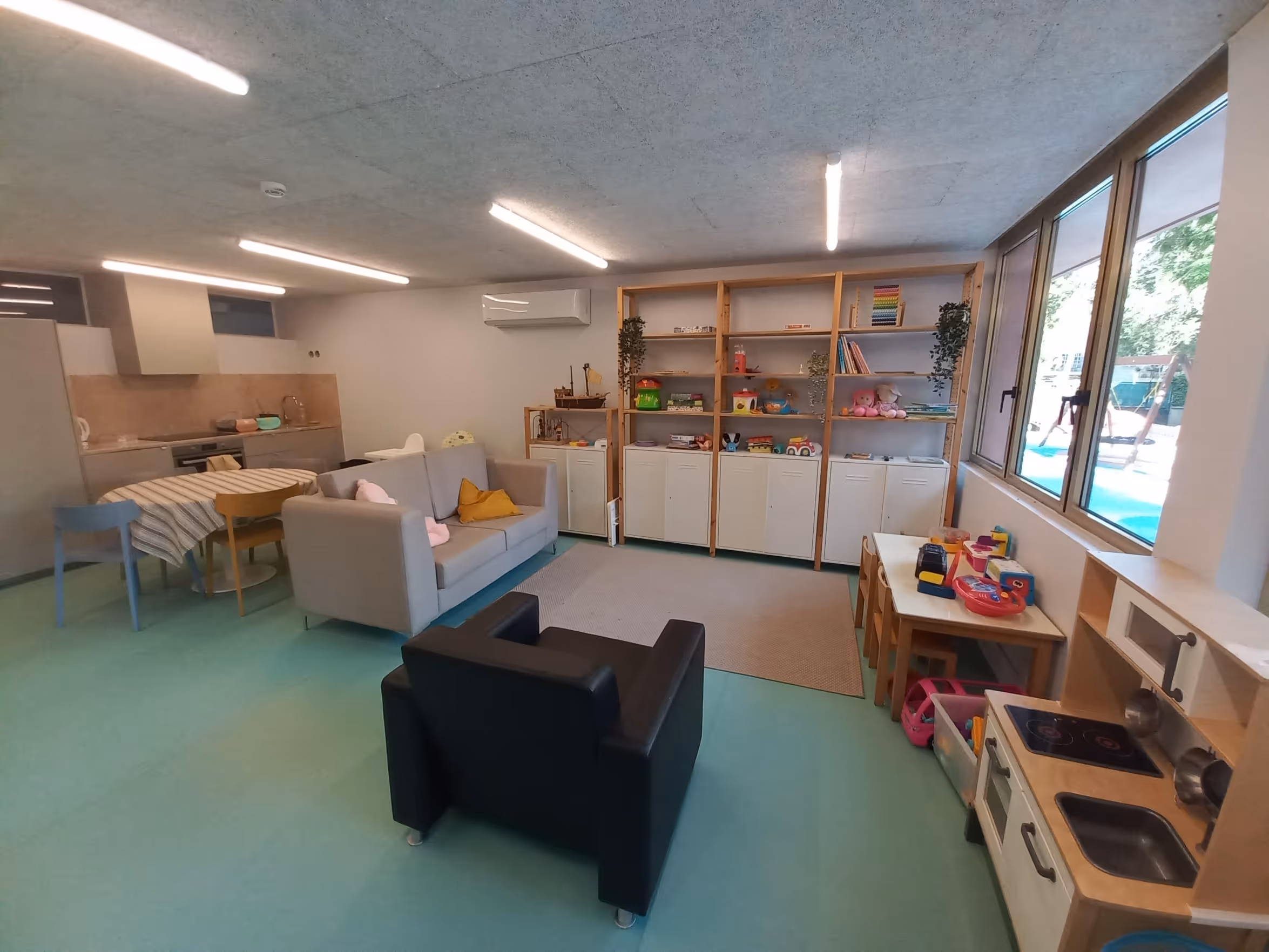 Bright playroom with toys, shelves, small tables, and seating near large windows and kitchenette.