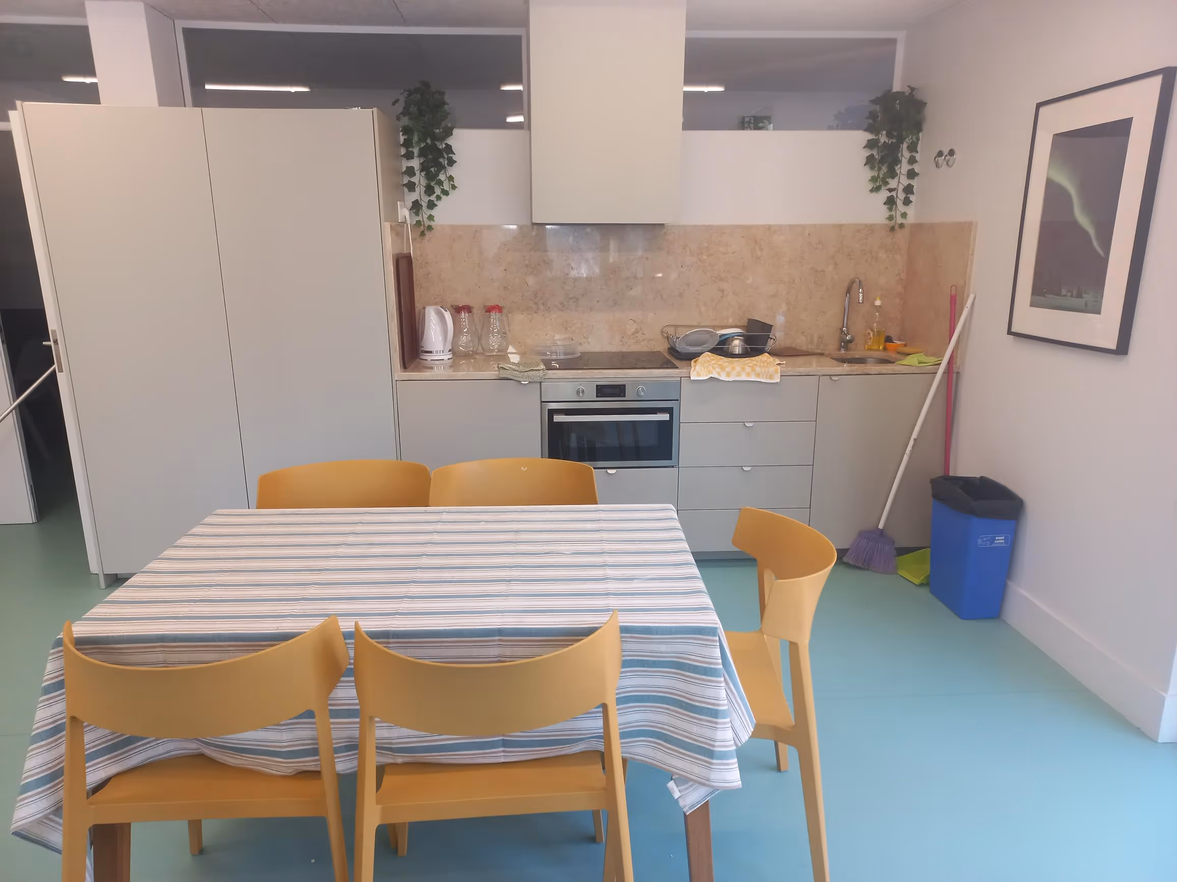 Simple kitchen with white cabinets, striped tablecloth on table, yellow chairs, cleaning tools, and wall art.