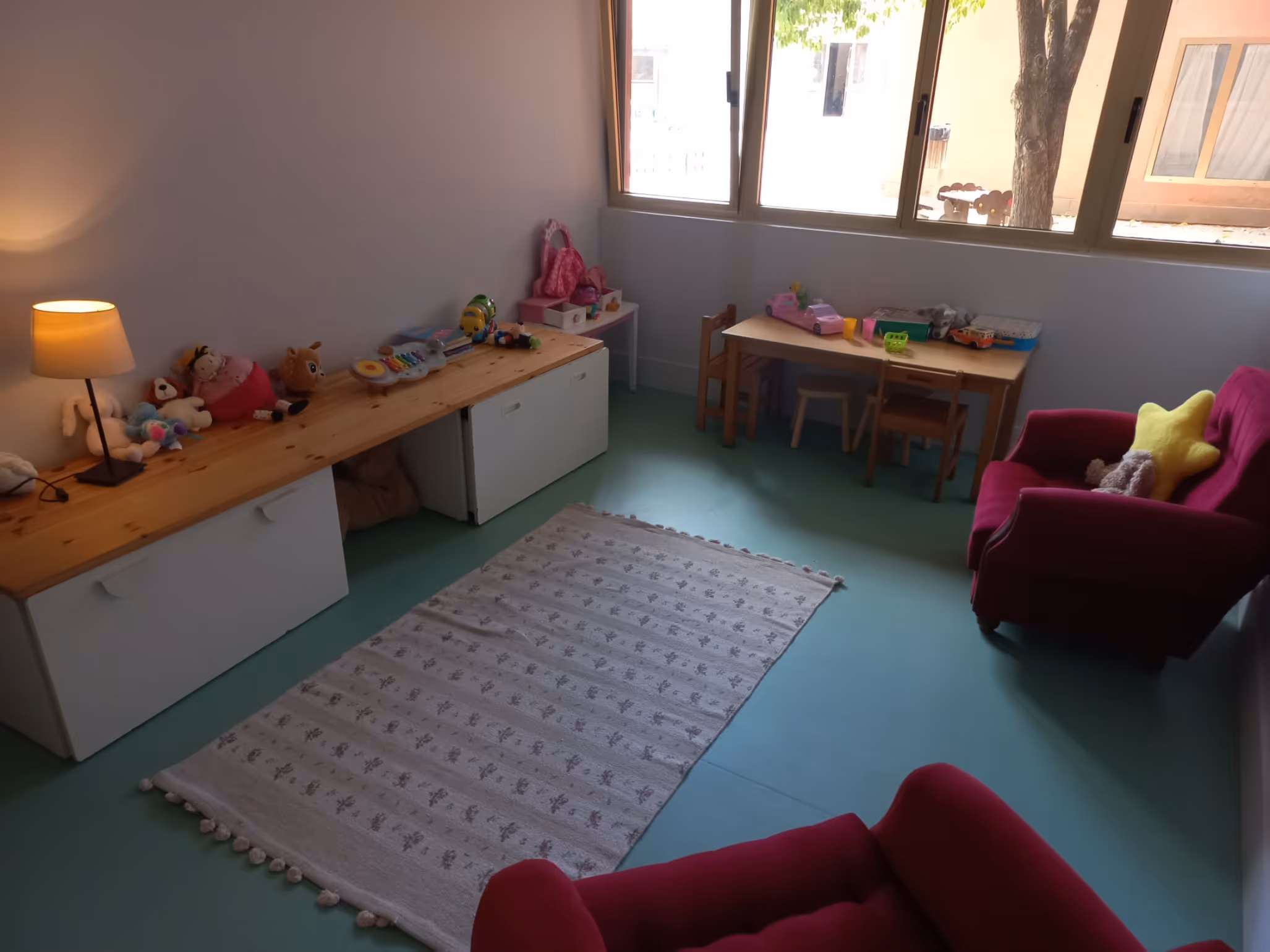 Children's playroom with toys on shelves, small table with chairs, and two red armchairs with cushions.