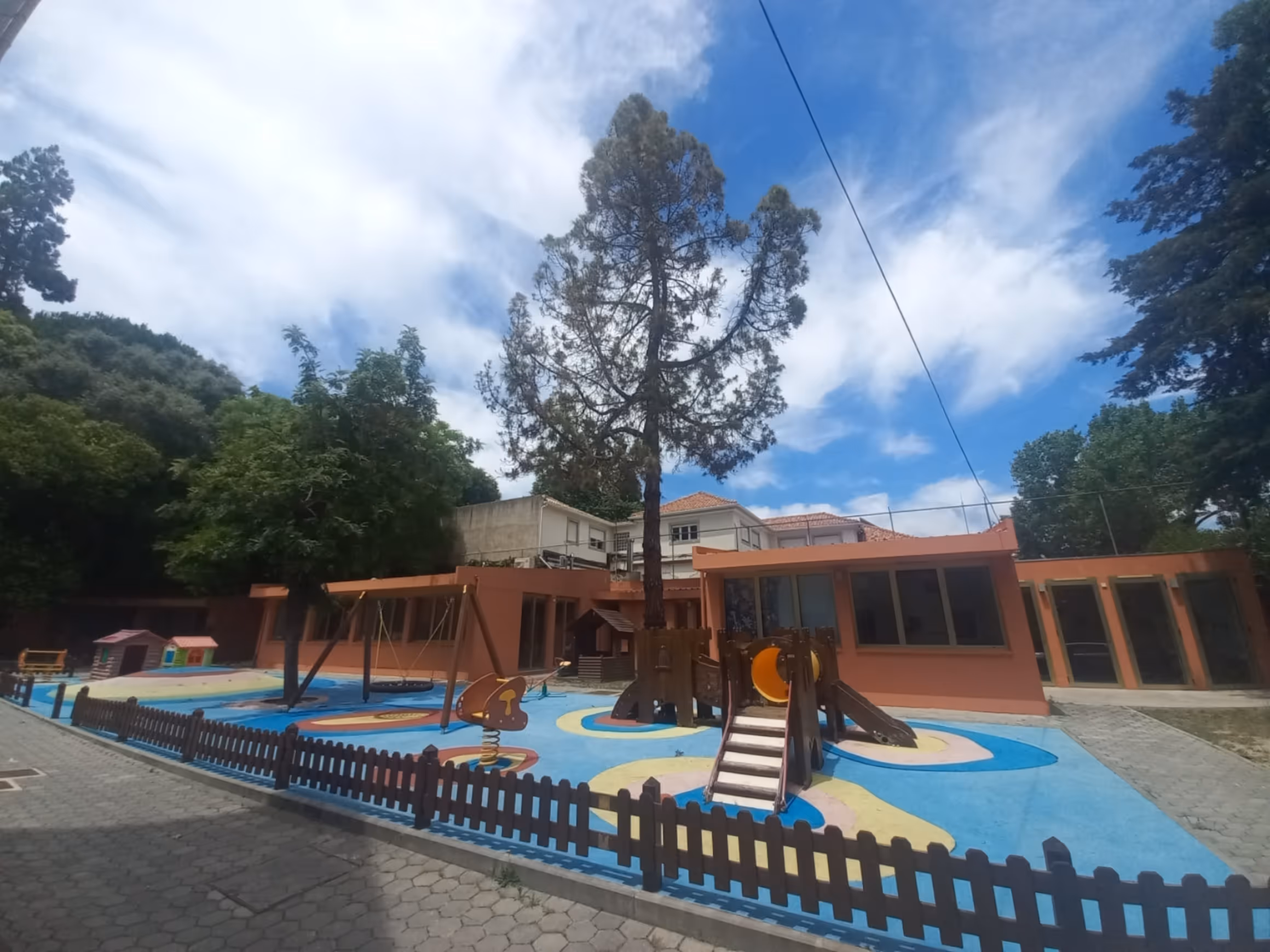 Outdoor playground with swings, slides, and playhouses surrounded by trees and a low fence.