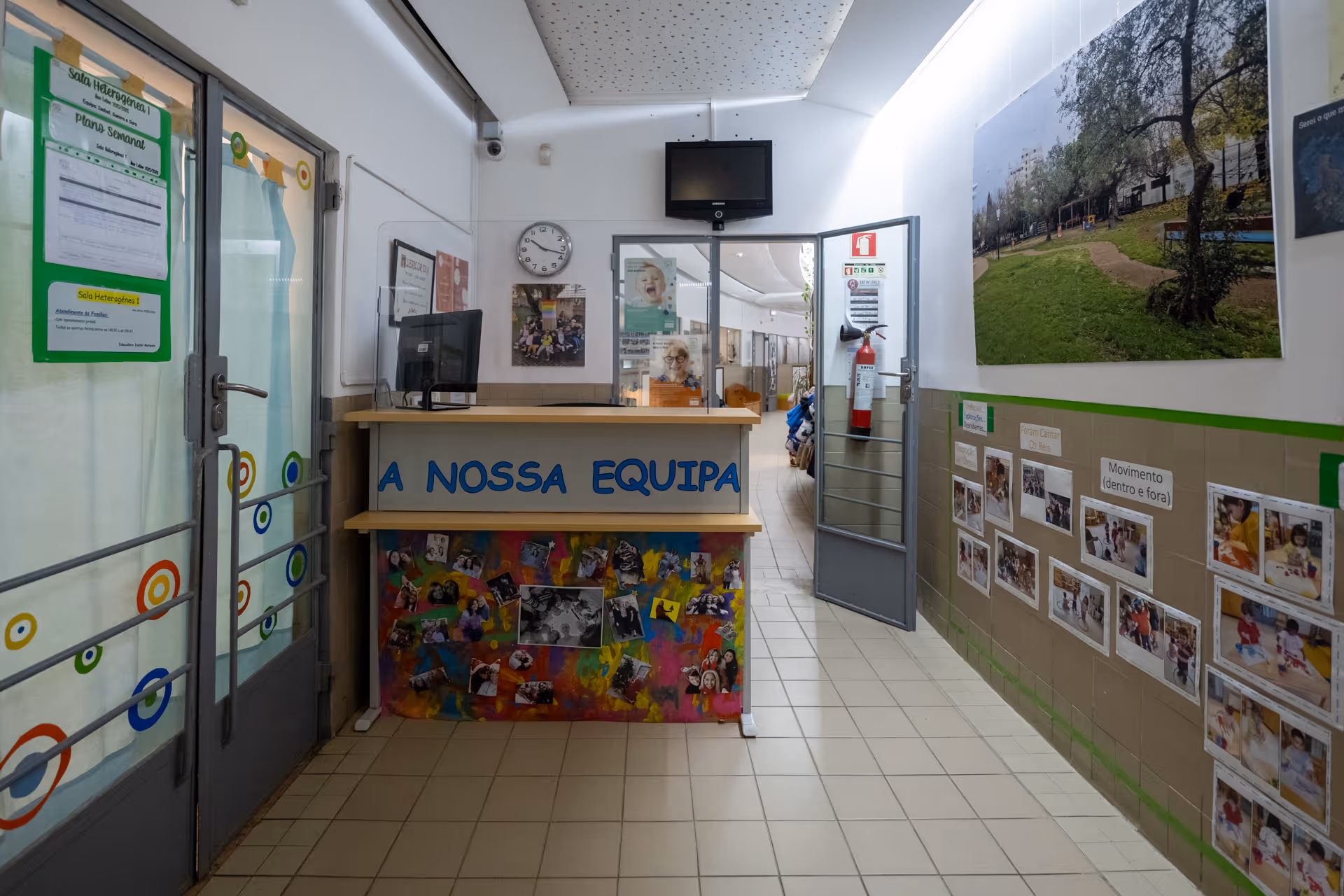 Reception area of a school with colorful desk labeled 'A NOSSA EQUIPA' and educational photos on walls.