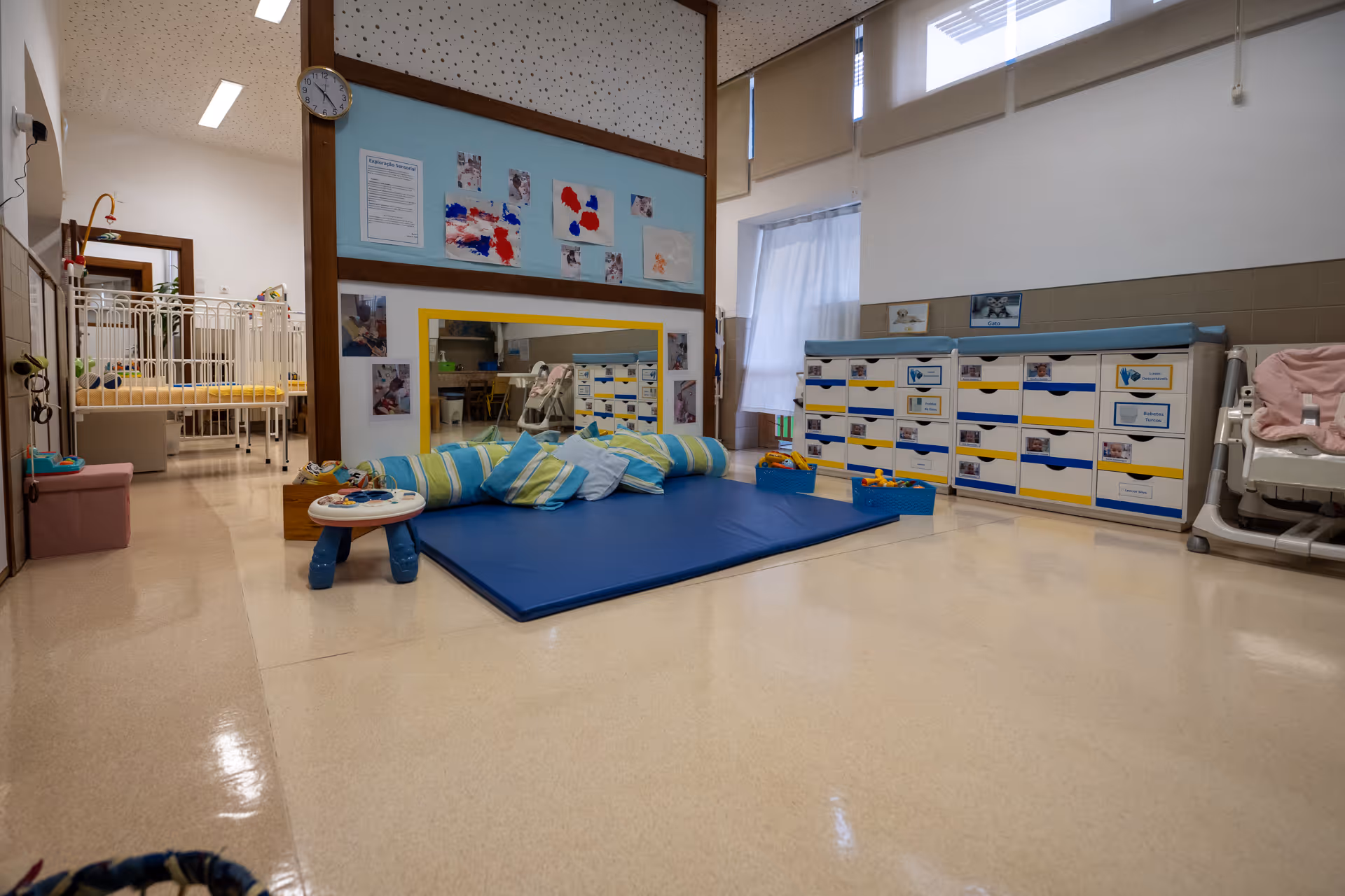 Bright daycare room with blue mat, striped pillows, toy baskets, storage drawers, and a small crib area.