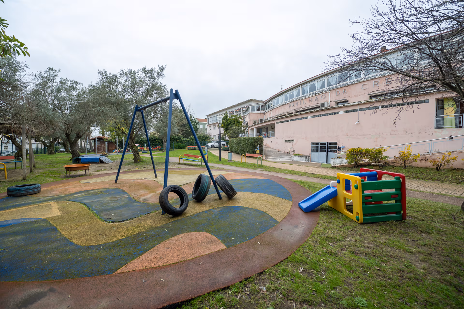 Outdoor playground with tire swings and small colorful slide beside a large pink building.