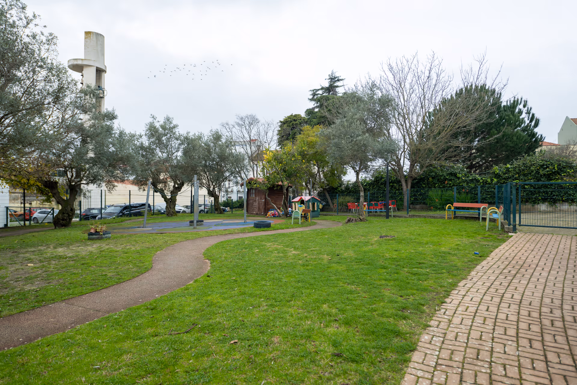 Park with green grass, playground equipment, benches, trees, and a paved walkway on an overcast day.