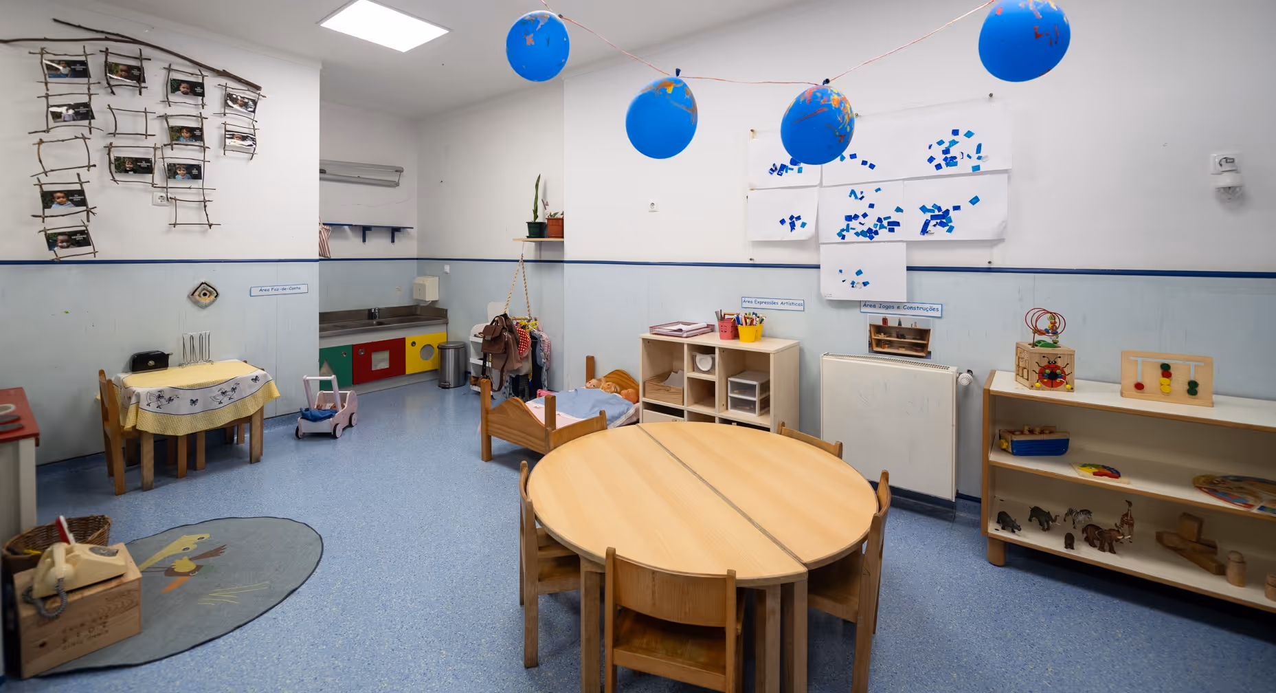 Empty preschool classroom with small tables, chairs, toys, blue floor, and balloons hanging from ceiling.