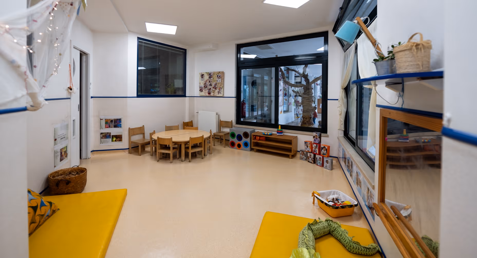 Bright preschool classroom with round table, wooden chairs, toys, mats, and a large window showing a tree decoration.