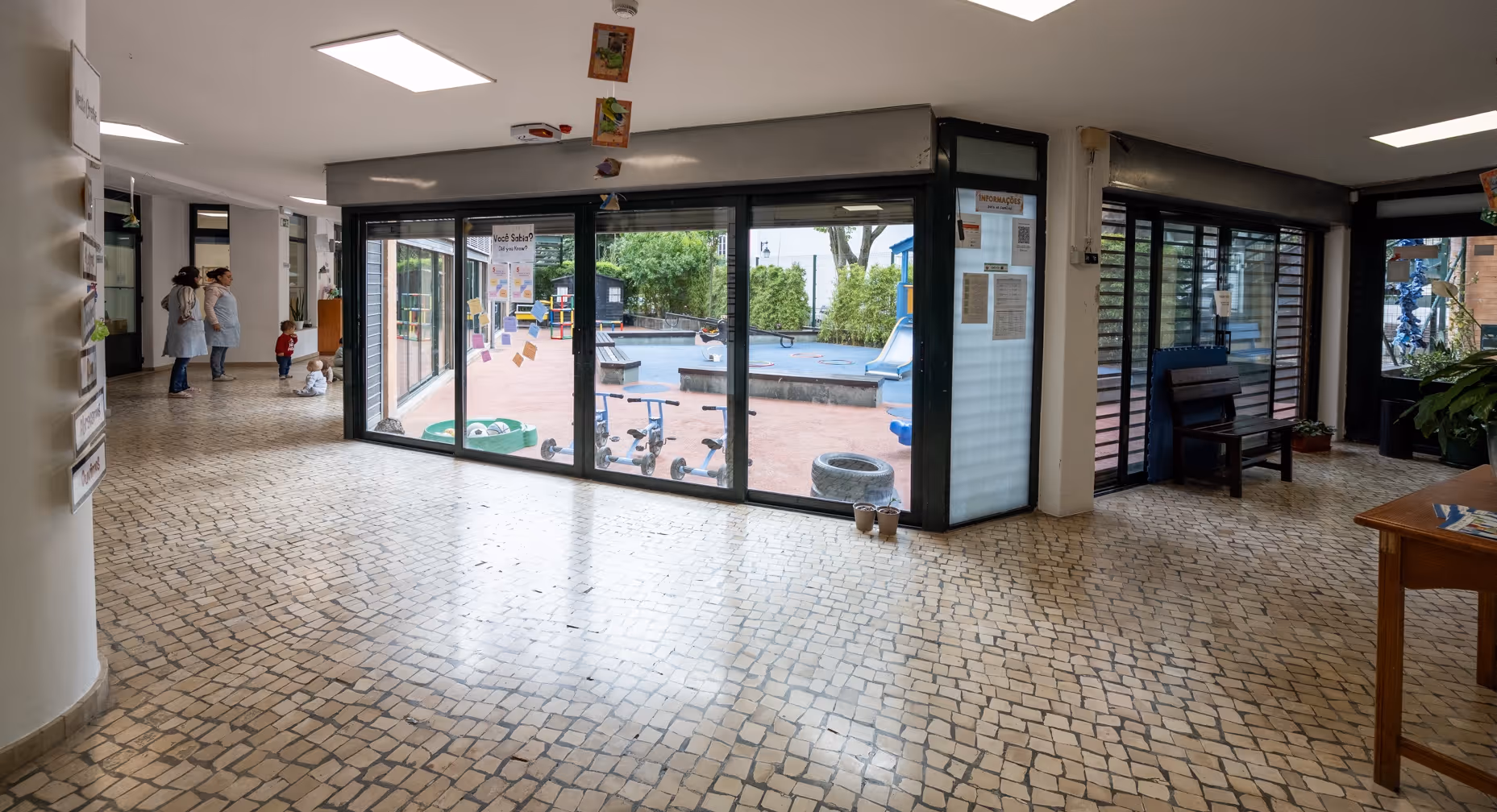 Indoor hallway with tiled floor, two women and two toddlers near large windows overlooking a playground.