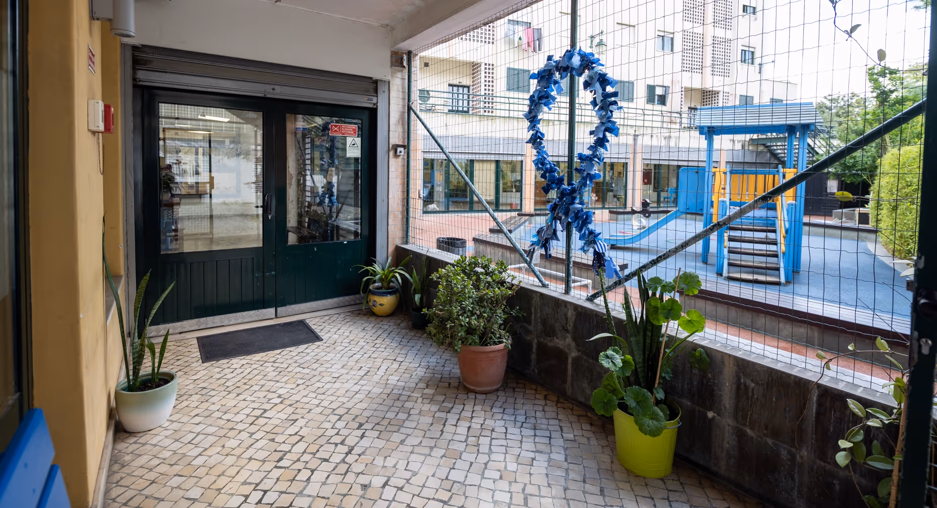 Entrance area with potted plants, tiled floor, and a netted railing overlooking a blue playground slide.